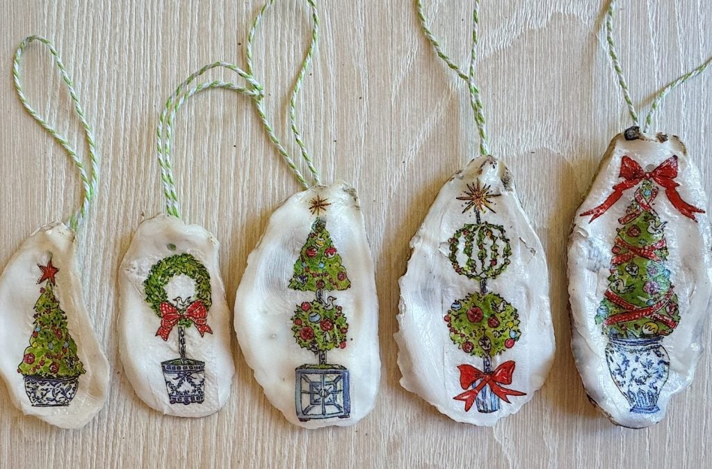 Five oyster shell ornaments with painted holiday designs, including trees and wreaths, attached to green and white striped strings.