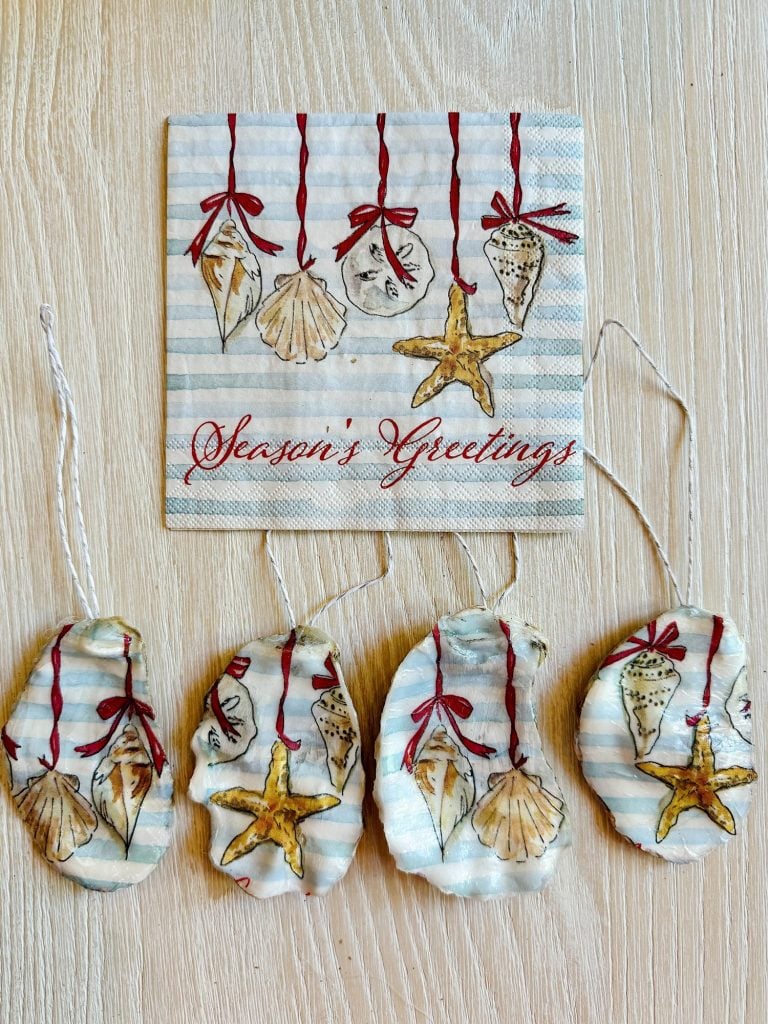 A napkin with seashell designs and "Season's Greetings" text, plus four seashell ornaments with red ribbon hangers on a light wooden surface.