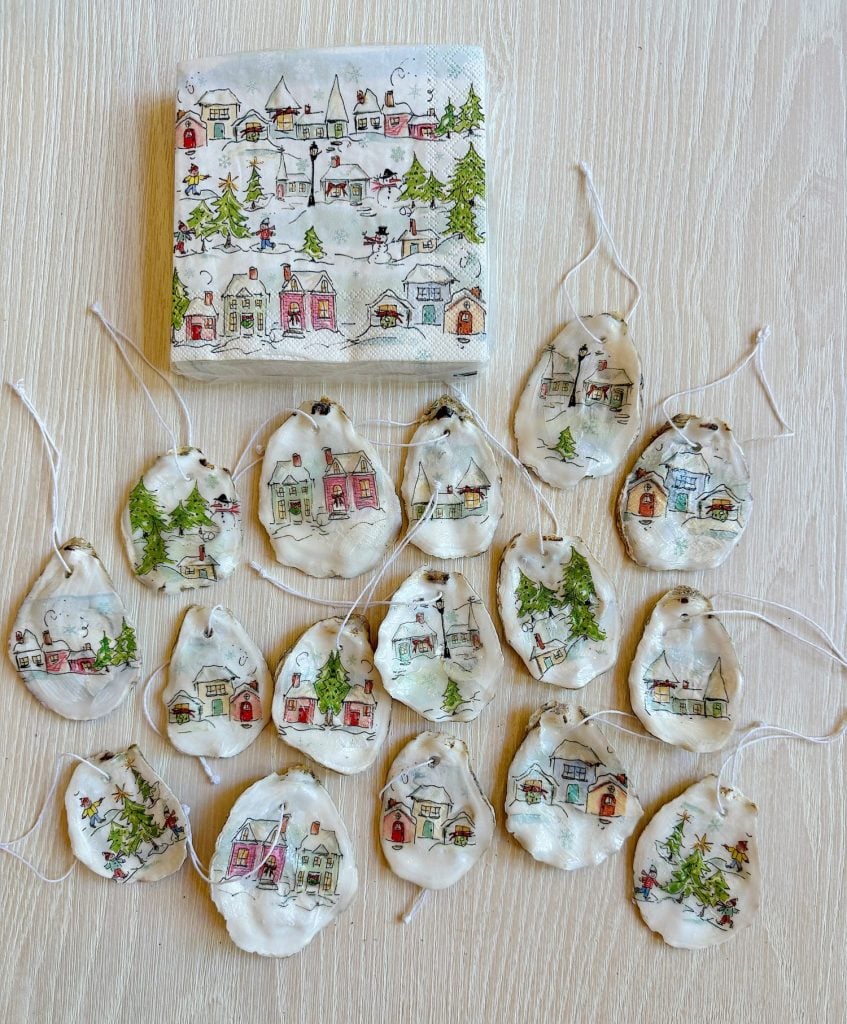 Handmade clay ornaments with painted winter village scenes are arranged on a light wood surface, alongside a matching napkin.