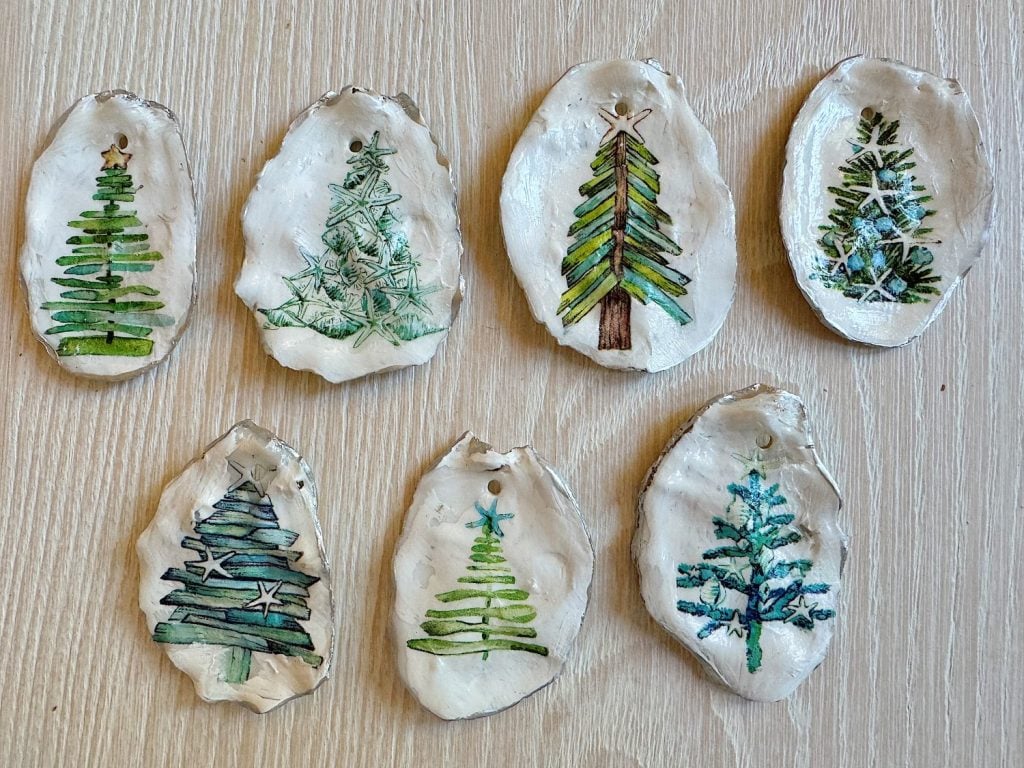 Seven painted oyster shells, each displaying a unique Christmas tree design in green and blue hues, arranged on a light wood surface.