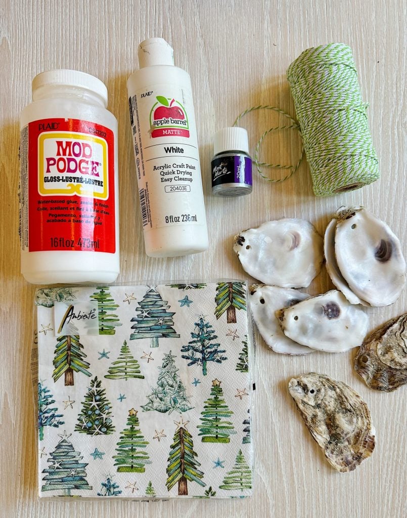 Craft supplies on a table: Mod Podge, white acrylic paint, green twine, a small purple paint bottle, decorative napkin with trees, and six oyster shells.