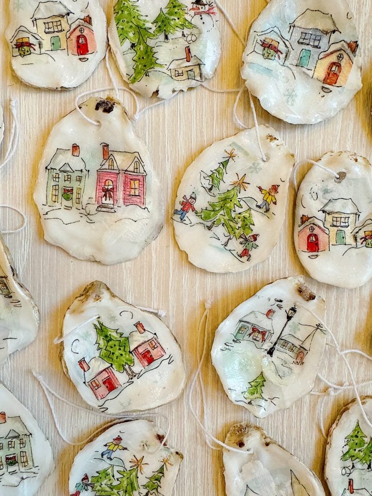 Hand-painted oyster shell ornaments showcase winter village scenes with charming houses and Christmas trees, elegantly arranged on a wooden surface.