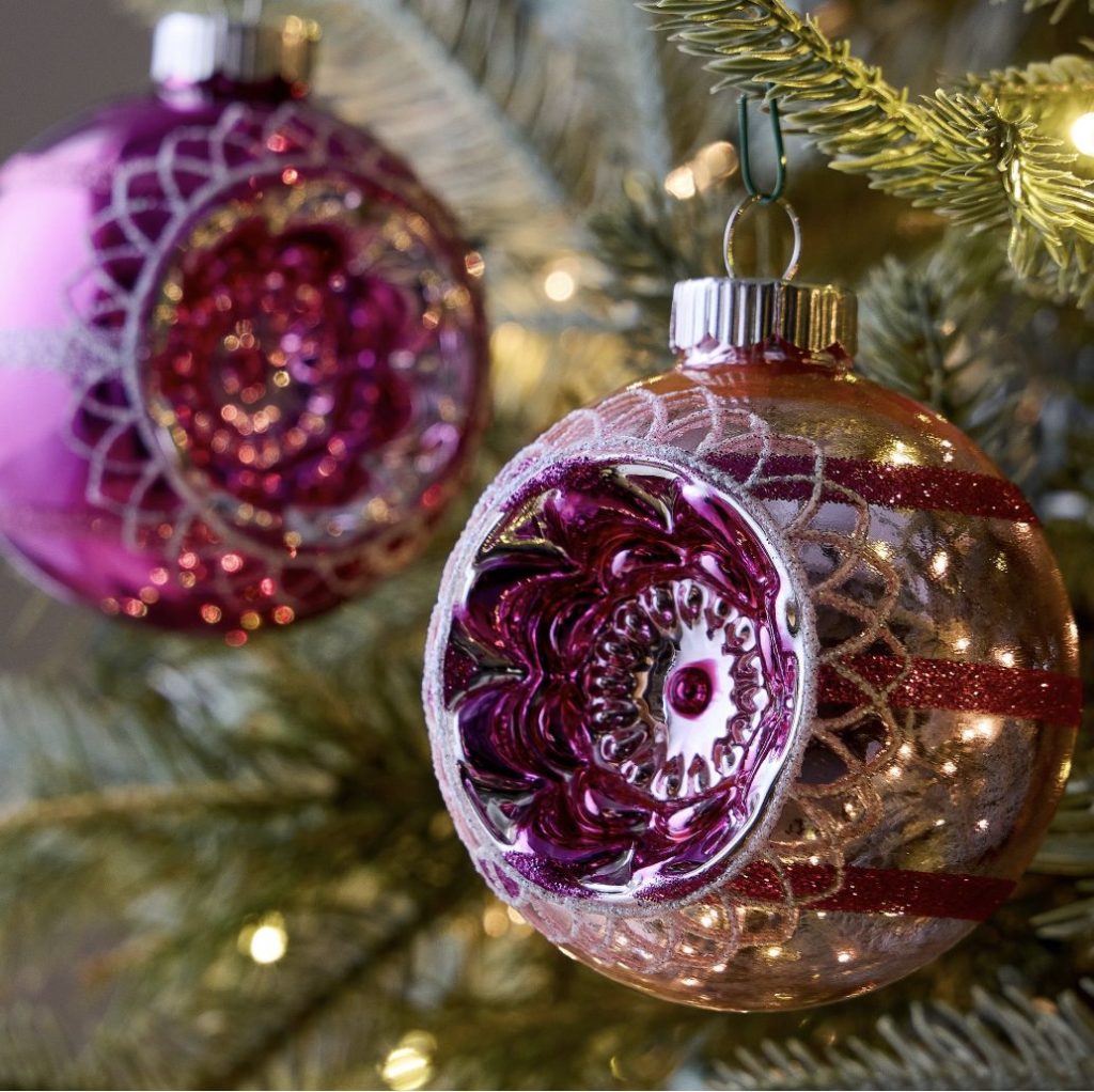 Close-up of two vintage purple and silver Christmas ornaments hanging on a tree, nestled among pine needles and glowing string lights.