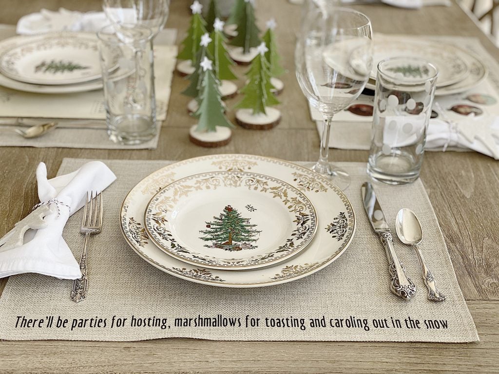 A holiday-themed dining table setting with plates featuring Christmas tree designs, surrounded by cutlery, glasses, and a napkin with a white holder, set on a placemat with festive text.