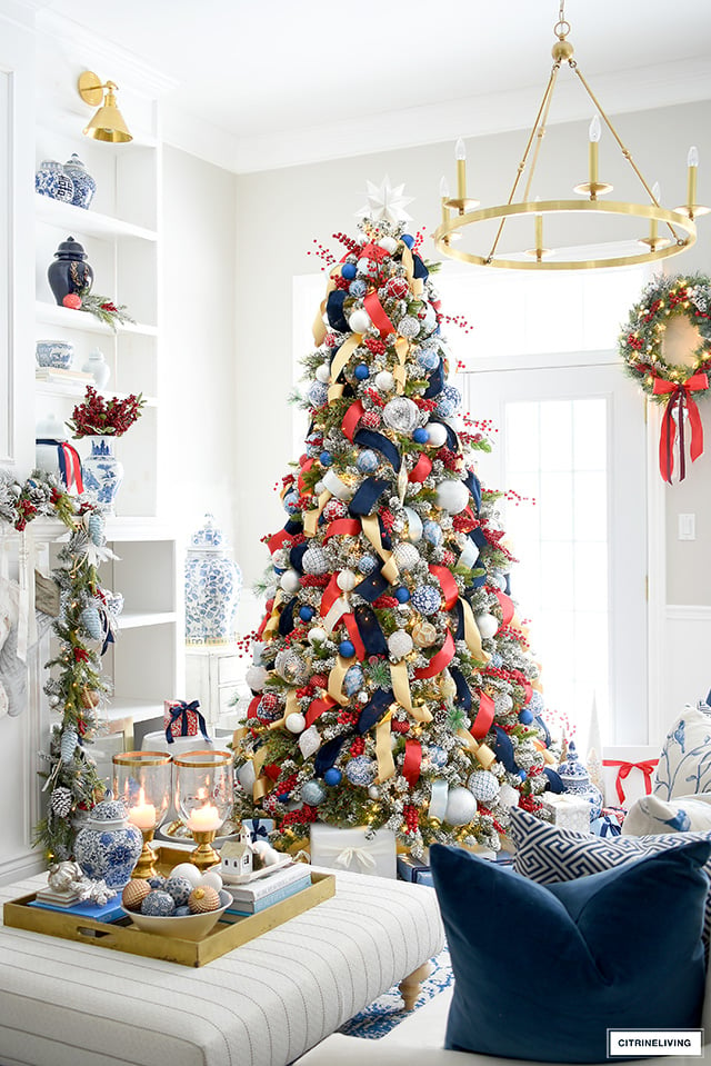 A brightly decorated Christmas tree with red, blue, and gold ornaments—the quintessential Christmas colors—stands in a living room. A wreath hangs on the door, and decorative items are tastefully arranged on a nearby table.