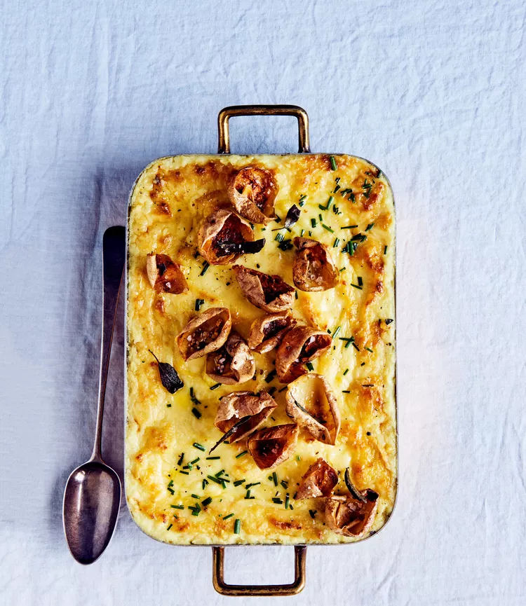 A baked dish with a golden crust, garnished with roasted garlic cloves and chives, sits on a white tablecloth. A spoon rests beside the dish.