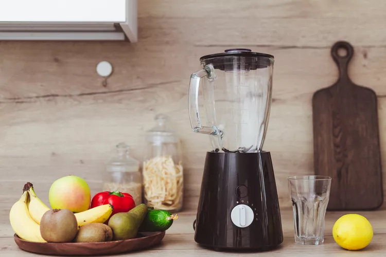 A blender on a kitchen counter next to a glass. A wooden bowl holds apples, bananas, kiwi, a pear, a cucumber, and a tomato. A cutting board and jars are in the background.