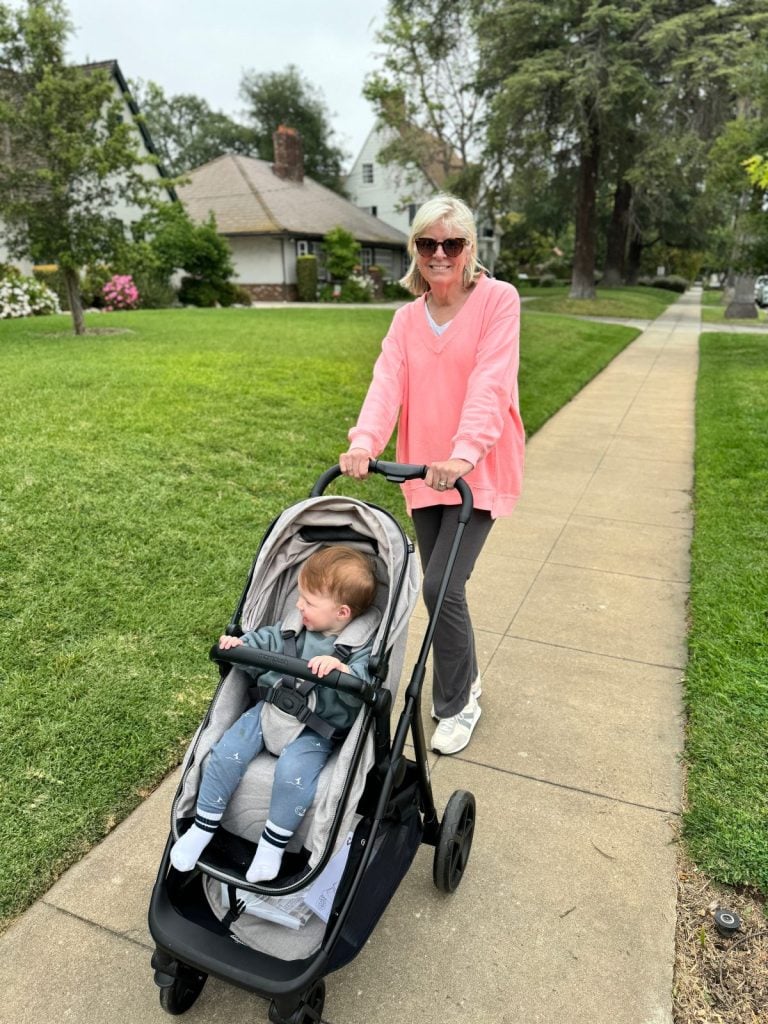 A woman in a pink sweater pushes a stroller with a seated child along a paved sidewalk in a suburban neighborhood.