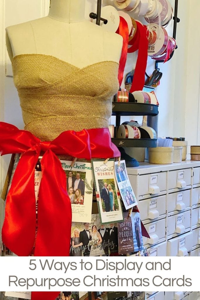 A mannequin wrapped in burlap and a red ribbon displays Christmas cards, with rolls of ribbon and storage drawers in the background.