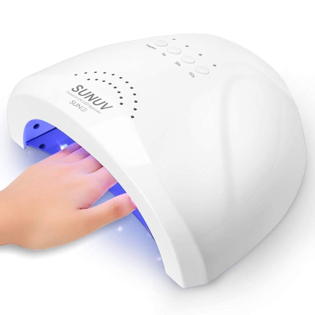 A hand is carefully placed under a white SUNUV LED nail lamp, its blue light gently curing both the nail polish and delicate clay ornaments waiting nearby.