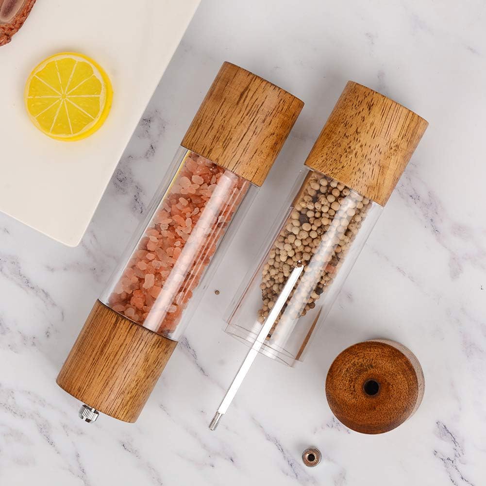 Two clear grinders with wooden tops and bottoms, filled with pink Himalayan salt and white peppercorns, rest on a marble surface next to a lemon slice—perfect for elevating your meals during the season of giving.