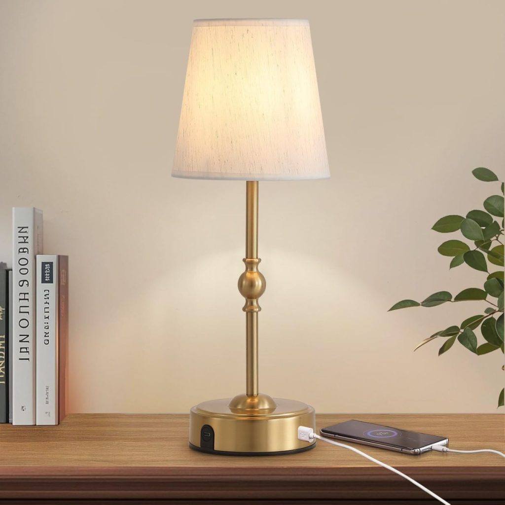 A brass table lamp with a white shade sits on a wooden surface next to books and a plant, while a phone charges via the lamp’s USB port—perfect lighting for cozy reading or setting the mood at your Christmas party.