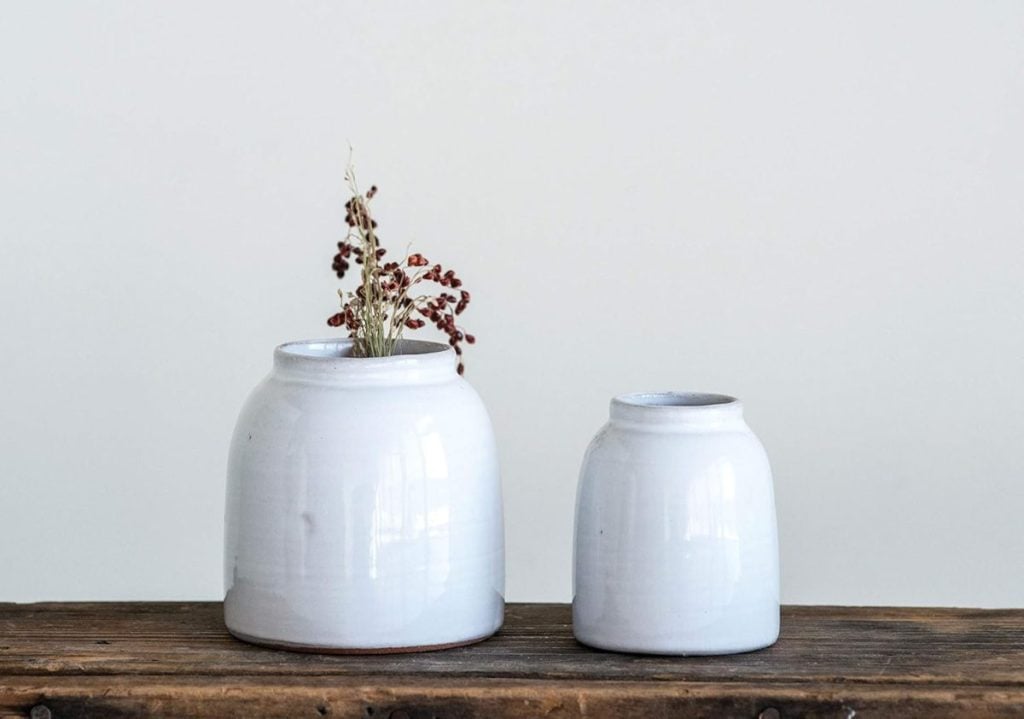 Two white ceramic jars rest on a wooden surface; the left jar holds dried red and beige flowers, evoking the cozy charm of a Christmas Morning Breakfast, while the right jar remains empty.