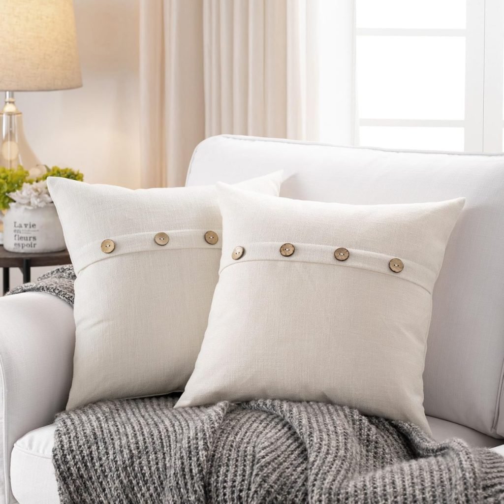 Two beige pillows with button accents are placed on a white sofa with a gray knitted throw. A lamp and small plant are in the background.