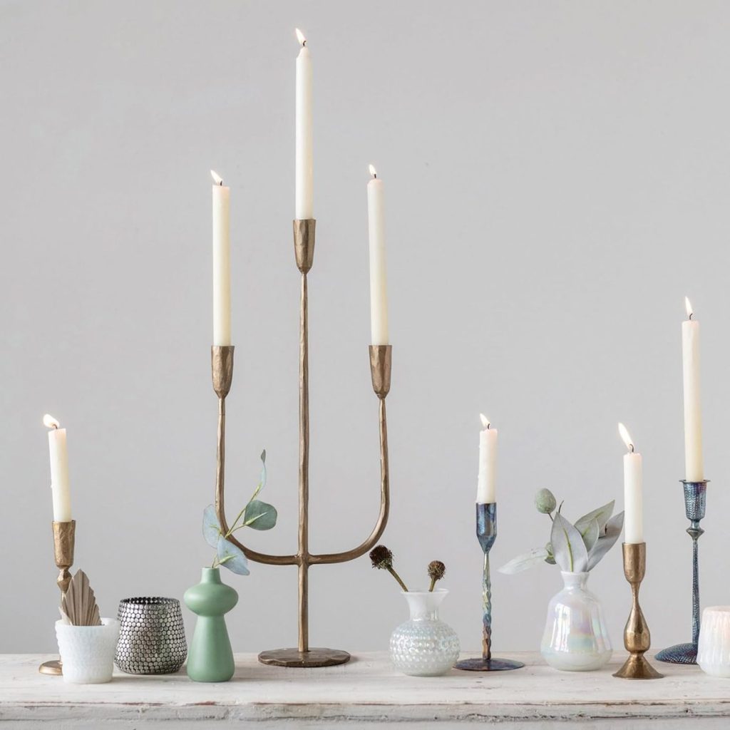 A group of lit candles in various holders, including a tall brass candelabra and assorted ceramic and glass vases, arranged on a light wooden surface against a plain background, evokes the warmth of the season of giving.
