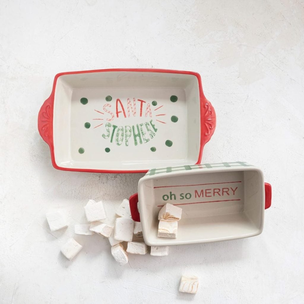 Two rectangular ceramic baking dishes, one reading "SANTA STOP HERE" and the other "oh so MERRY," are perfect for your Christmas party, with marshmallows scattered beside them on a white surface.