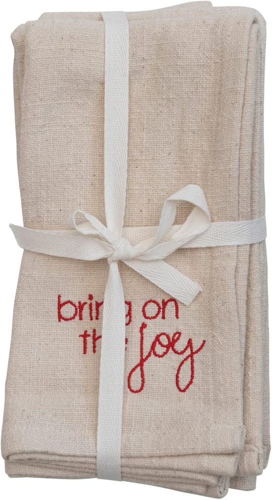 A folded beige cloth napkin tied with a white ribbon, featuring the phrase "bring on the joy" embroidered in red thread, makes a charming accent alongside festive clay ornaments.
