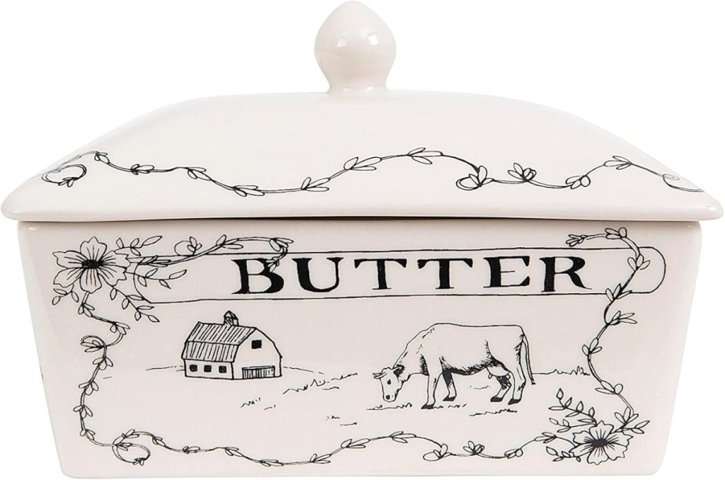 A white ceramic butter dish with a lid, decorated with black line drawings of a cow, a barn, flowers, and vines, and labeled "BUTTER" on the front—perfect for the season of giving.