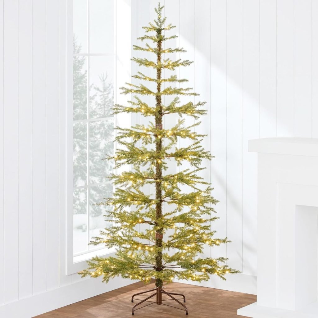 A slim artificial Christmas tree with sparse branches and white lights stands in a bright room with white walls and a large window.