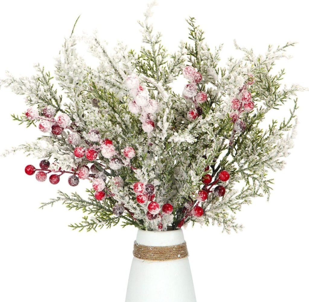 A white vase holds frosted evergreen branches with red berries, creating a festive display.