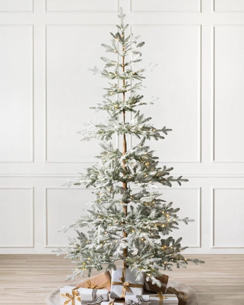 A tall, snow-dusted Christmas tree with sparse branches is decorated with white lights and placed in front of a white paneled wall. Several wrapped gifts are beneath the tree.