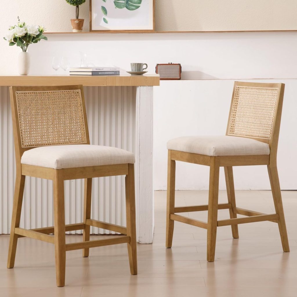 Two wooden bar stools with rattan backs and cushioned seats are placed near a kitchen counter with decor and a painting above.