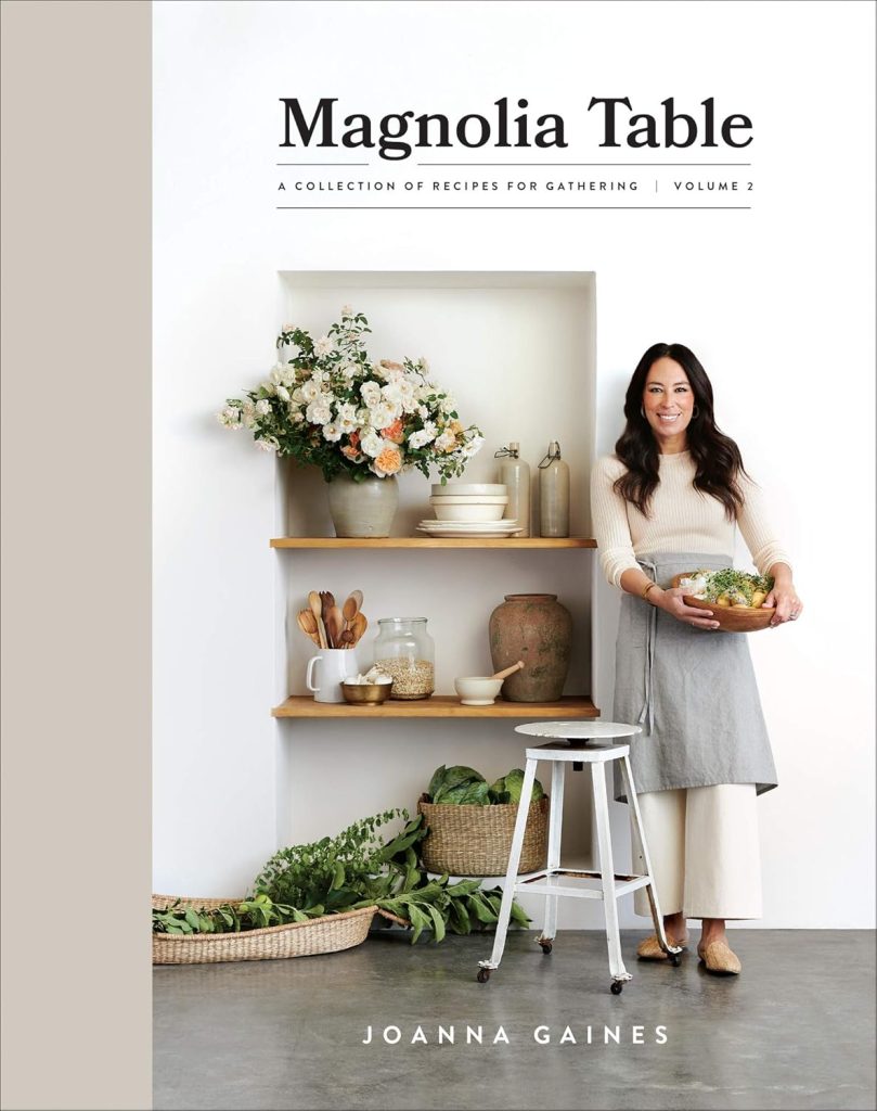 A woman stands beside wooden shelves with dishes and flowers, holding a bowl of salad—a perfect scene for the season of giving. The book cover reads "Magnolia Table Volume 2" by Joanna Gaines.