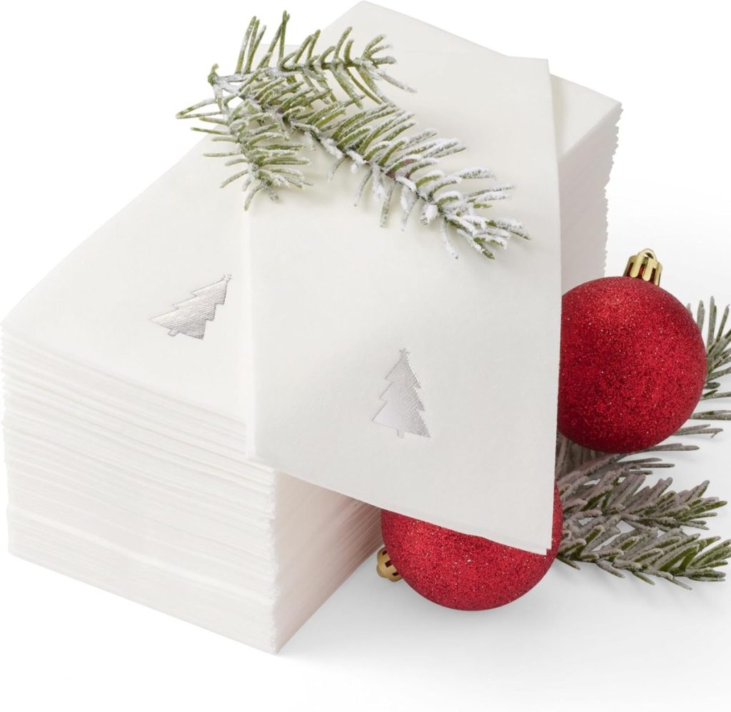 A stack of white napkins with silver tree imprints, adorned with red ornaments and fir branches.