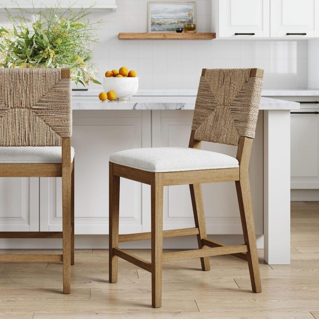 Two wooden bar stools with woven backs and cushioned seats are positioned in a white kitchen. A bowl of oranges and a potted plant are on the marble countertop.