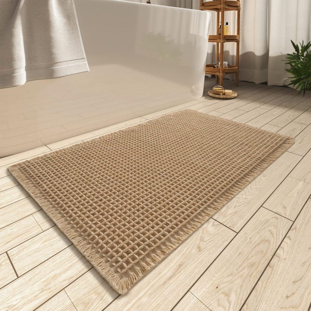 A beige bath mat with a waffle texture on a wooden floor next to a white bathtub.