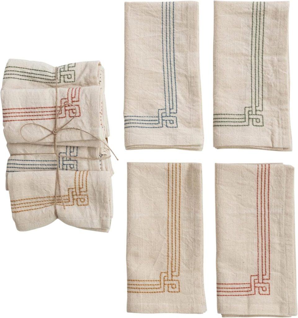 Four folded linen napkins with colored embroidered borders in green, blue, red, and yellow—plus an additional set tied with string—make a thoughtful touch for the season of giving, beautifully displayed on a white background.