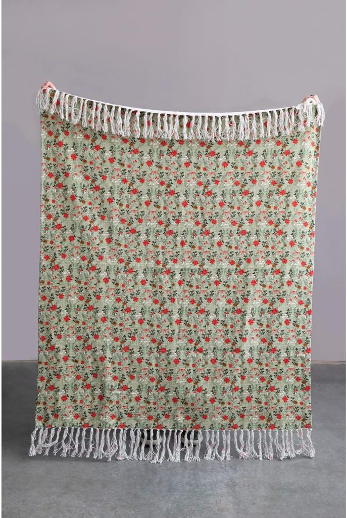 A person holds up a rectangular, fringed blanket with a green background and a red and black floral pattern—perfect for cozying up during the season of giving—against a plain gray backdrop.