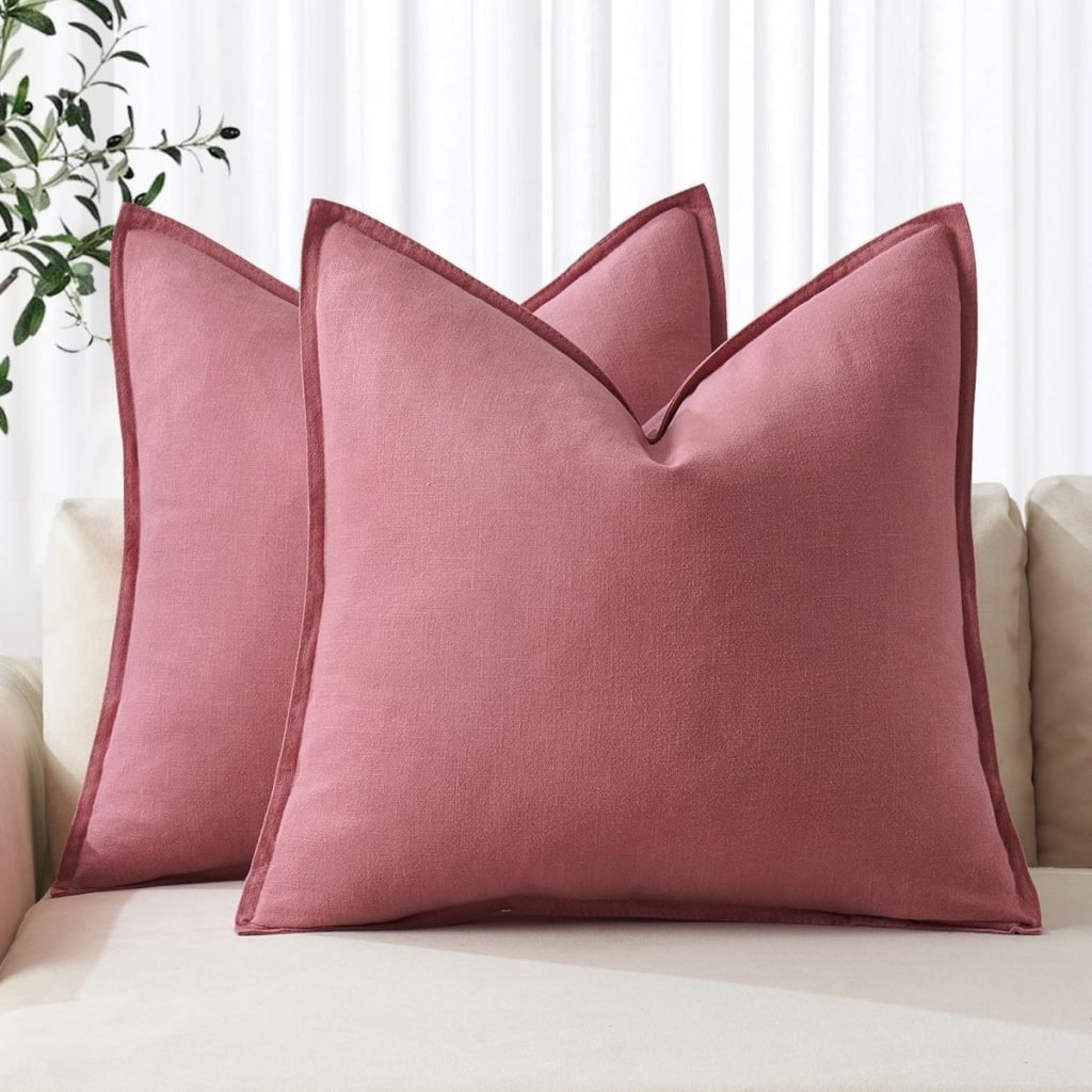 Two large pink pillows with pointed corners are placed on a beige sofa. A plant with green leaves is visible in the background.