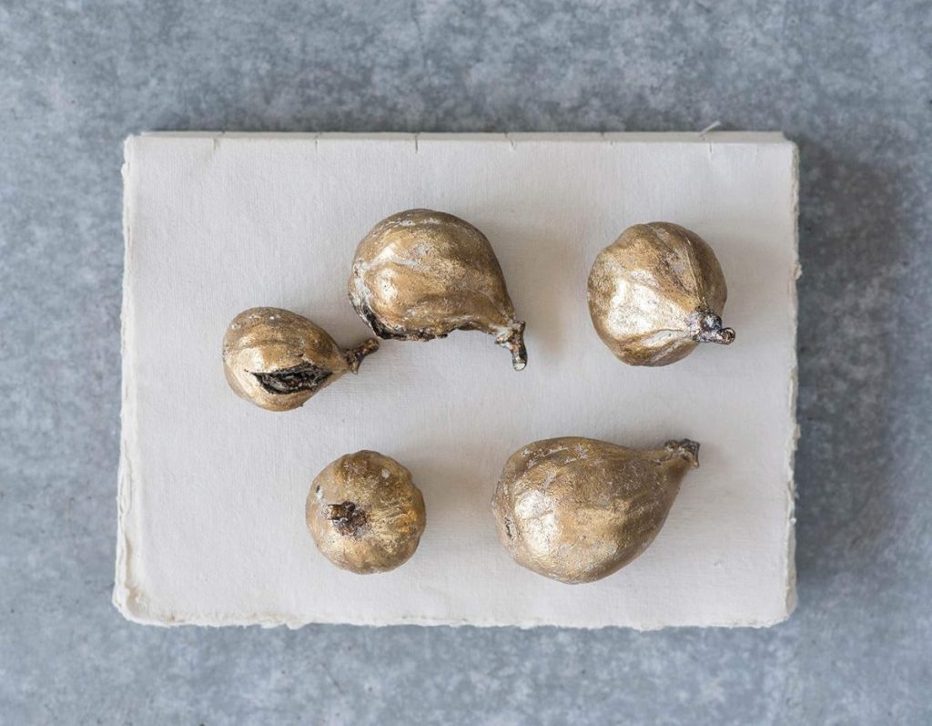 Five gold-painted dried figs, perfect for the season of giving, are displayed on a rectangular white slab against a gray textured background.