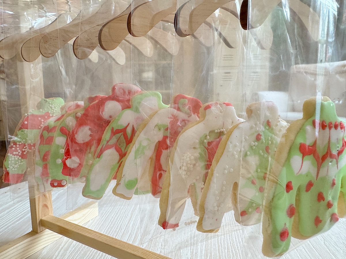 Colorful sweater-shaped cookies with holiday-themed icing hang on wooden hangers inside clear bags.