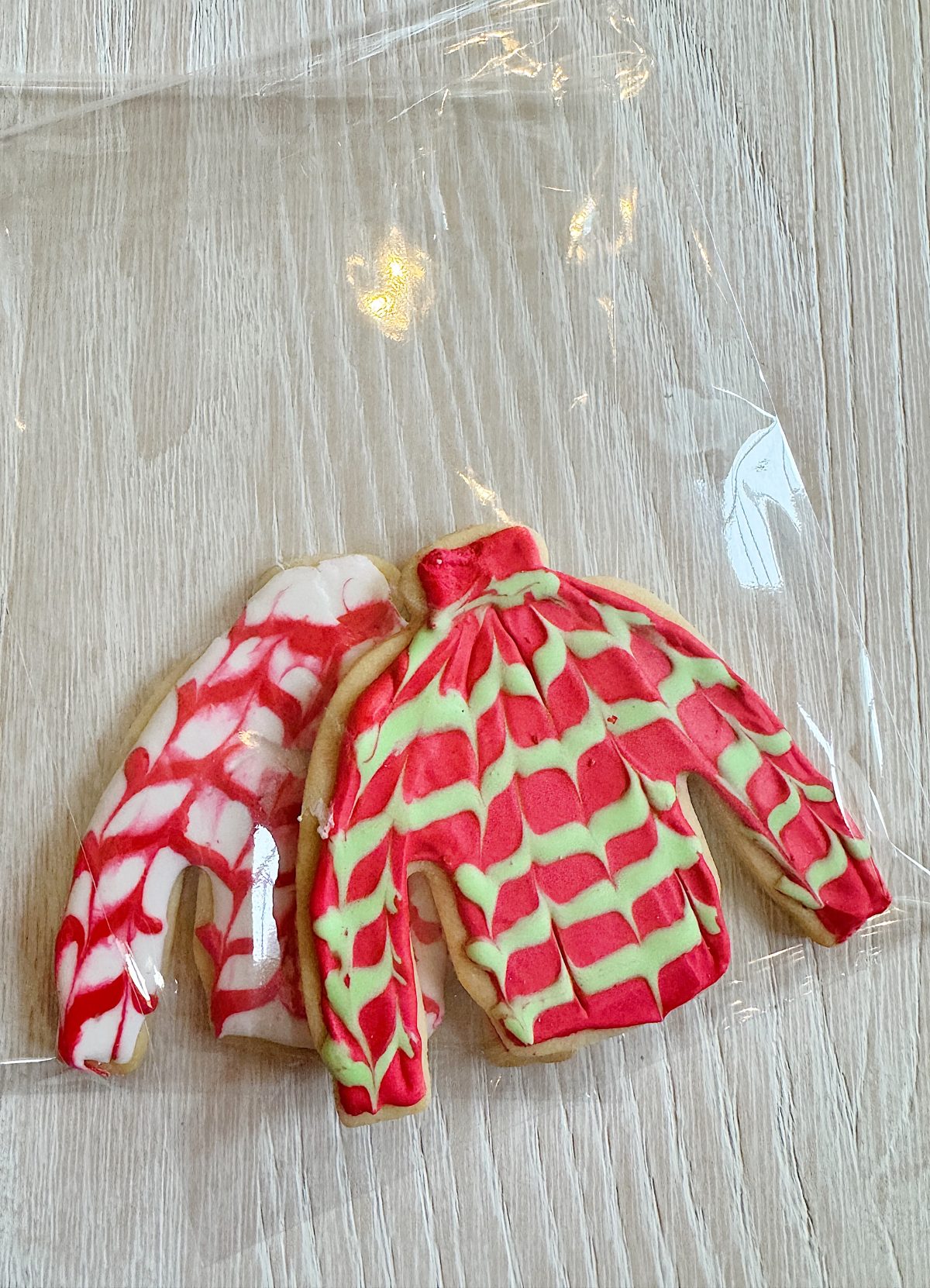 Two sweater-shaped cookies decorated with red and green icing patterns on a clear plastic wrap placed on a wooden surface.