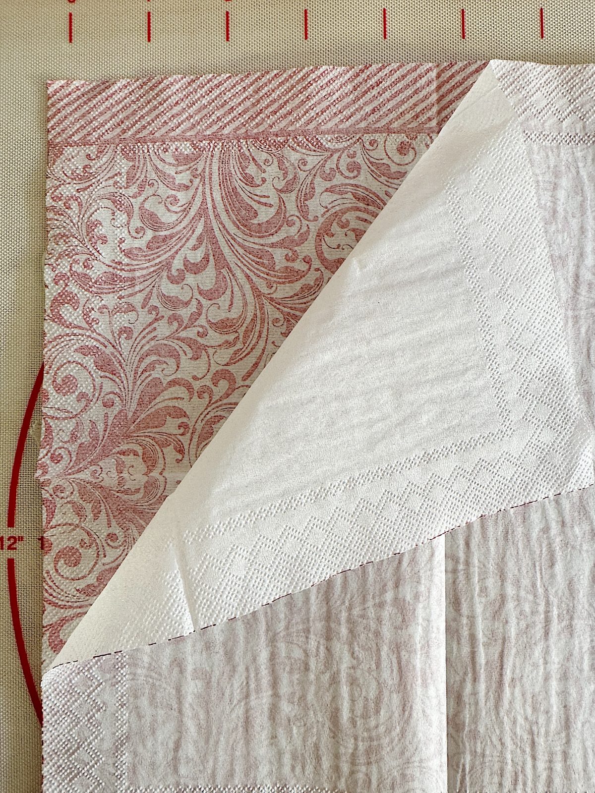 A white paper napkin is partially unfolded, revealing its patterned underside with a red floral and swirl design. A measuring grid is in the background.