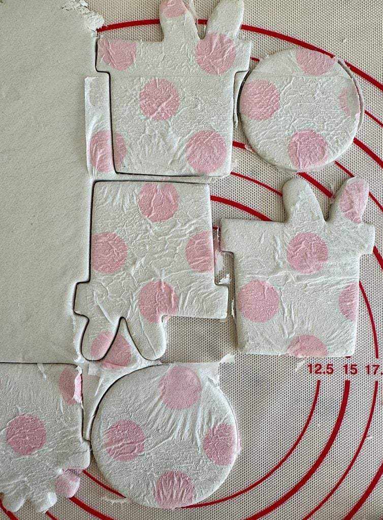 Cut-out pieces of white and pink polka-dotted dough shaped like bunnies and circles on a silicone mat with a red circular pattern.