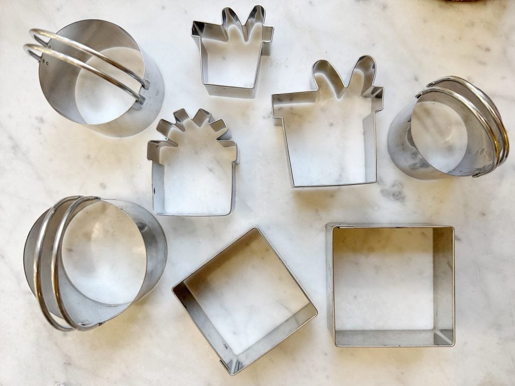 A collection of metal cookie cutters in various shapes, including gift boxes and circles, displayed on a marble surface.