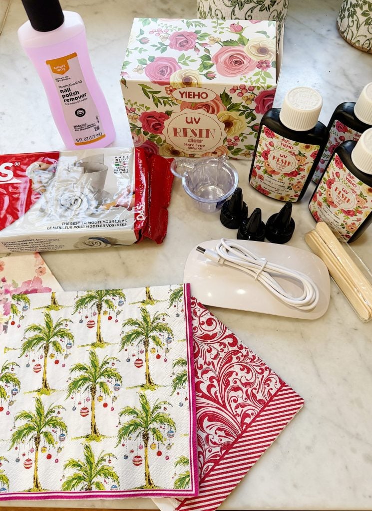 Crafting supplies on a table, including resin kits, palm tree and floral napkins, wooden stir sticks, nail polish remover, and a white device with USB cable.