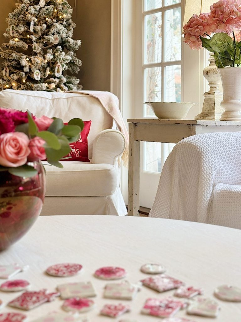 Cozy living room with white armchairs, a decorated Christmas tree, pink flowers, and a table with pink and white cookies.