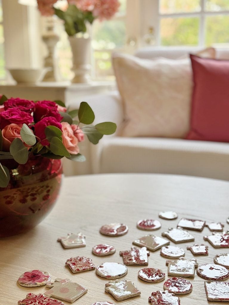 A table with assorted decorated cookies in floral shapes and colors, a bowl of roses nearby, and a sofa with pink cushions in the background.