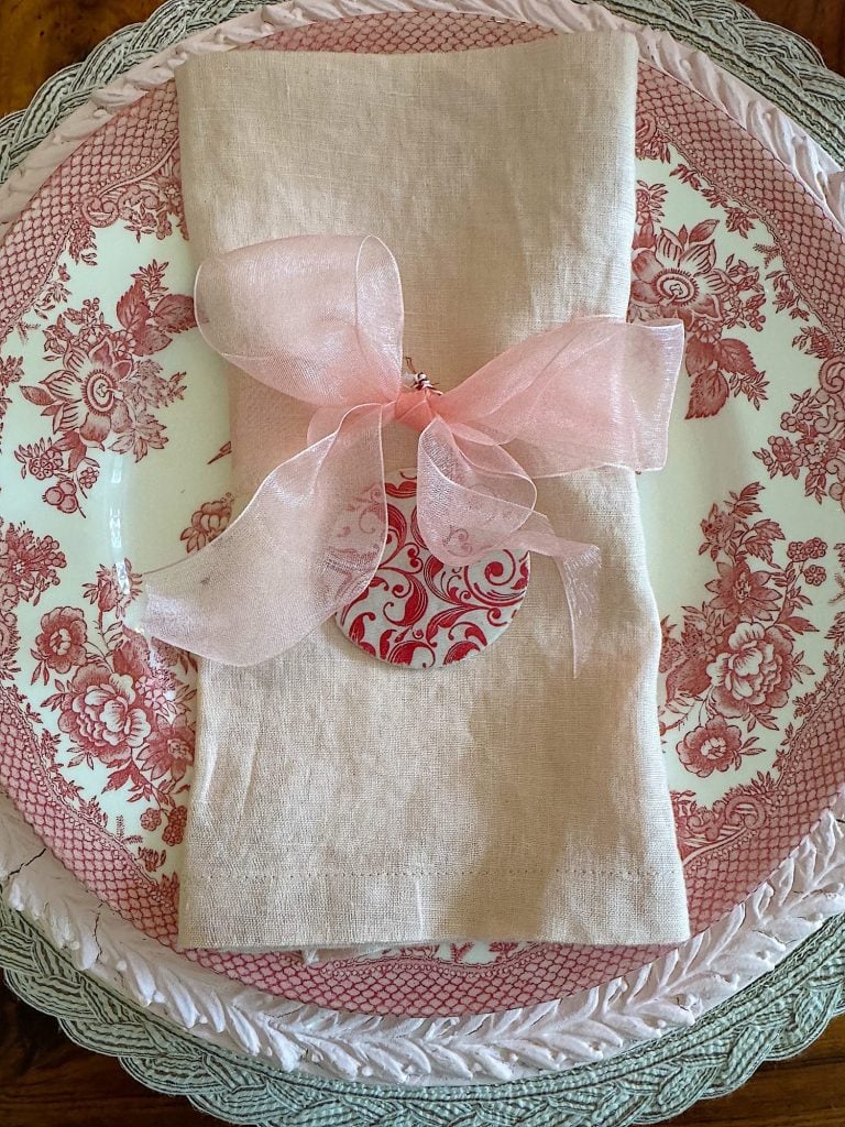 A pink napkin with a decorative pink ribbon and ornament is placed on a red and white floral plate.