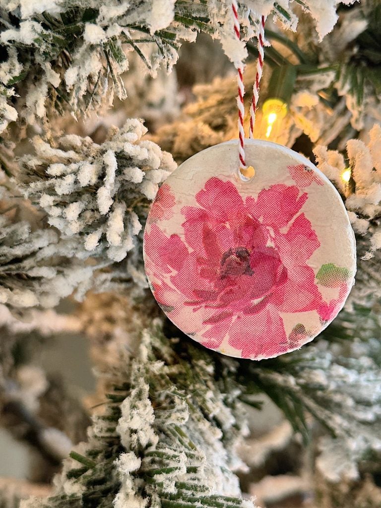 A floral ornament with a pink flower design hangs on a snowy Christmas tree branch adorned with lights.