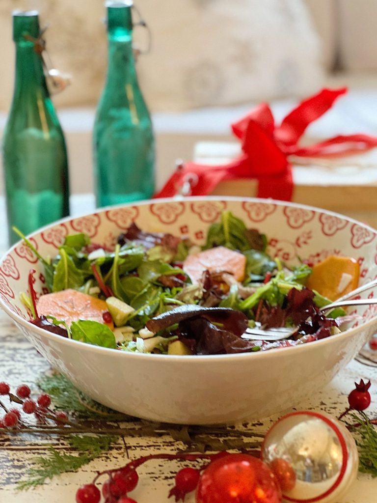 A festive salad with mixed greens and grapefruit in a decorative bowl, surrounded by red ornaments and green glass bottles.