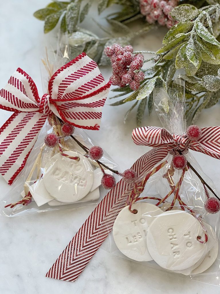 Two clear gift bags tied with red and white striped ribbons, containing white clay tags with names. Decorated with red berries and greenery, the bags rest on a marble surface.