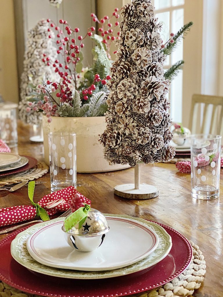 Festive dining table with a pinecone tree centerpiece, red and white accents, dotted glasses, and decorative plates.