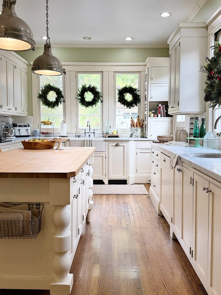 A bright kitchen with white cabinets, a wooden center island, hanging lamps, and wreaths on windows.