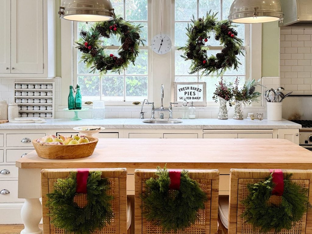 A decorated kitchen features a wooden table with wreaths on the chairs, two hanging lights, and three wreaths in the windows. A "Fresh Baked Pies" sign sits by the sink.