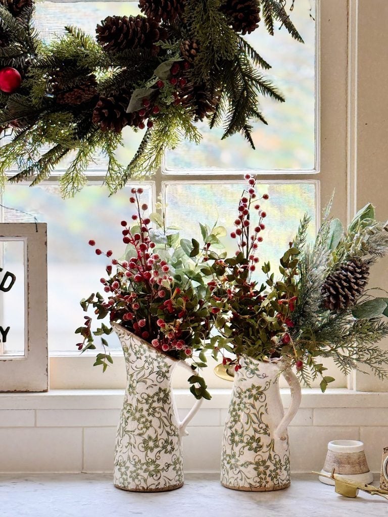 Two floral pitchers with red berries and greenery sit on a windowsill, beneath a pinecone and evergreen garland.